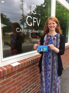 Office manager is standing in front of the store window, smiling, and holding the breastfeeding welcome here logo cling, which is a stylized picture of nursing dyad in front of the shape of NC