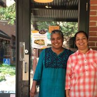 Two women are standing in front of Vimala's Curryblossom Café, smiling into the camera.