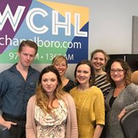 Six employees of WCHL, five women and one man, smiling into the camera with the WCHL logo behind them.