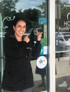 Dr. Holman is standing in front of the glass door to the business, smiling and pointing at the breastfeeding welcome here logo, which is a stylized picture of nursing dyad in front of the shape of NC.