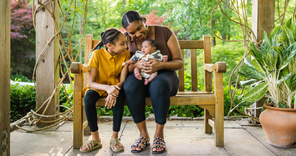Mother holding baby and sitting next to older child on a bench.