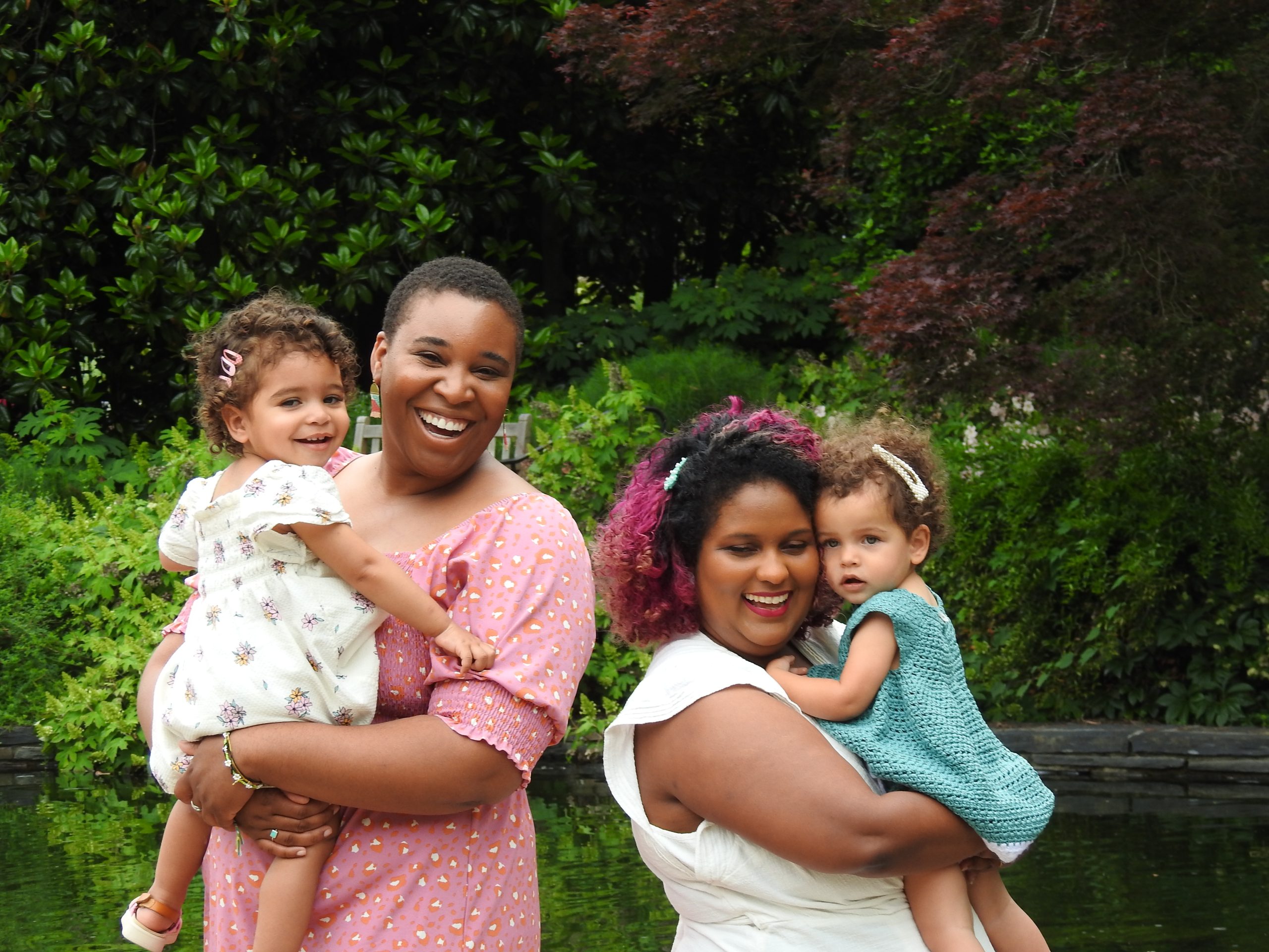Two mothers laughing and holding their toddler daughters in a garden