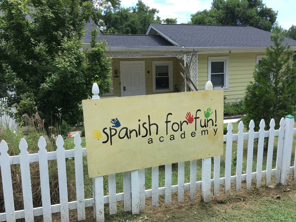 Single story building with a door and two windows; a sign with the words "Spanish for Fun academy" is on the picket fence that is in front of the building.
